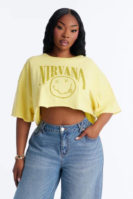 Woman in an oversized yellow Nirvana cropped t-shirt with a green smiley logo, paired with light blue baggy jeans. White background.