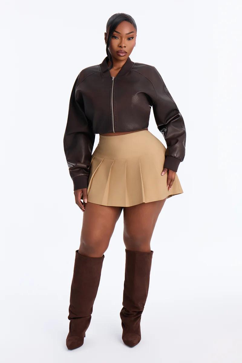 Woman wearing a dark brown cropped leather jacket, beige pleated mini skirt, and brown knee-high boots.