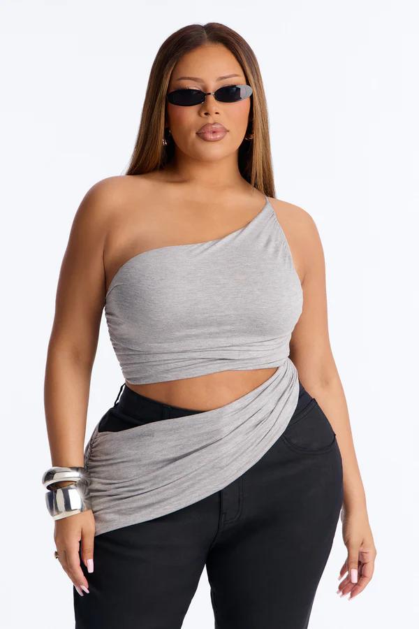 Woman in a grey one-shoulder crop top with a midriff cutout, black pants, black narrow sunglasses, and a silver cuff bracelet.