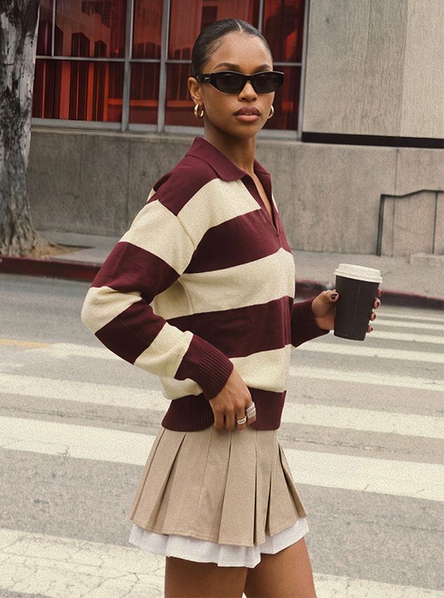 Mujer con suéter polo de rayas granate y minifalda plisada beige, que luce un estilo preppy urbano elegante para la moda de otoño.
