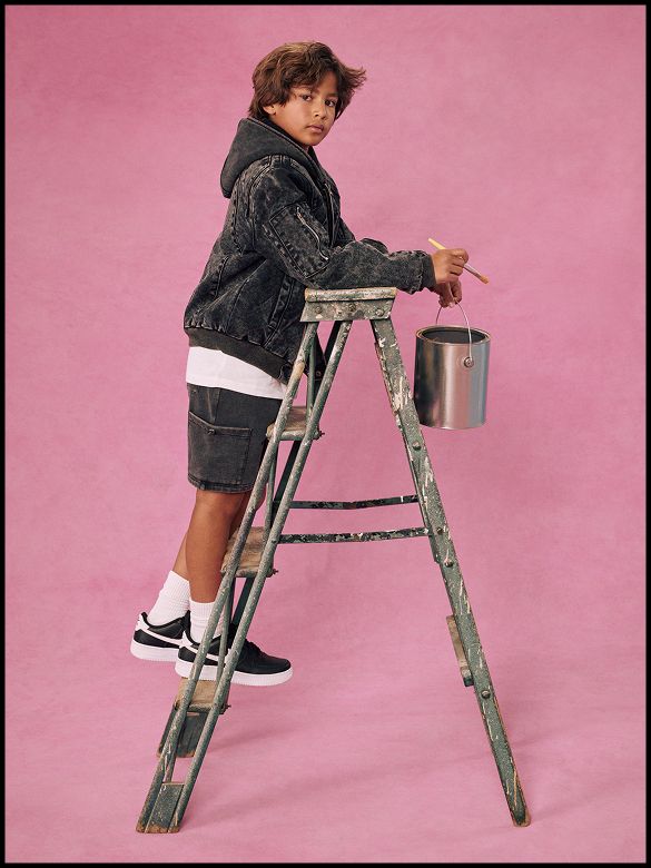 Boy in black denim jacket stands on a ladder with a paint can against a pink background, showcasing a trendy kids streetwear fashion collection.