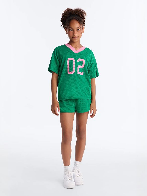 Young girl modeling a green mesh jersey top with pink trim and "02," matching shorts, and sneakers. New comfy kids' sportswear collection.