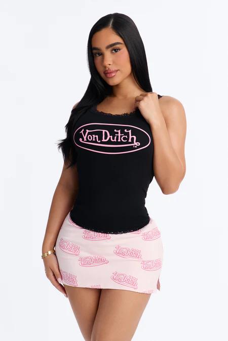 Woman in a black Von Dutch tank top with pink trim and logo, paired with a light pink patterned mini skirt, looking at camera.