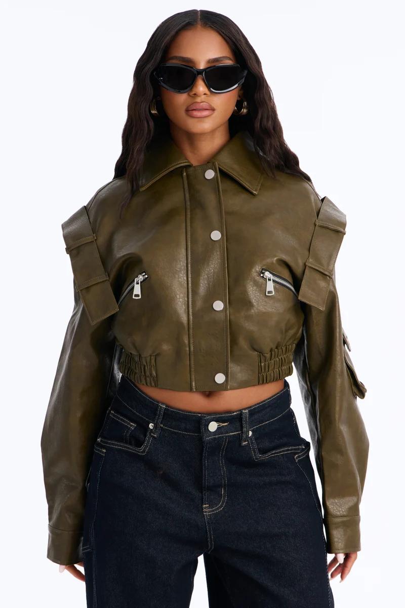 Woman wearing a cropped olive green faux leather utility jacket with silver zippers and dark blue jeans.