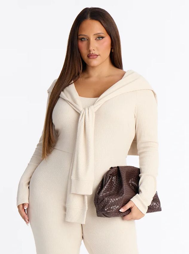 Plus-size woman wearing a cream ribbed jumpsuit with a tied sweater over her shoulders, holding a brown woven clutch bag.