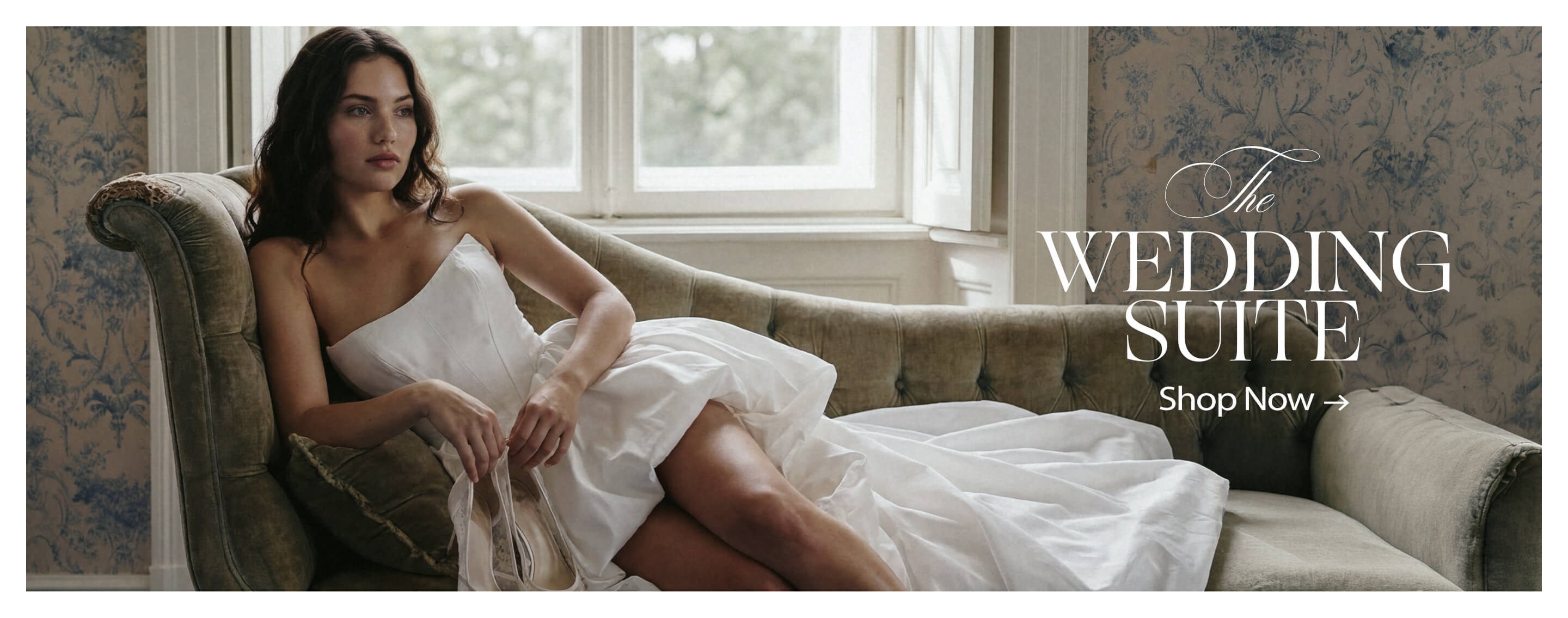 Woman in white top on olive couch, holding elegant pearl bridal heels. Promotes The Wedding Suite collection. Shop bridal shoes now.