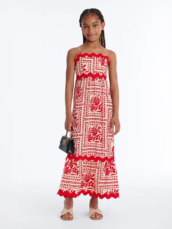 Smiling girl in a cream and red floral maxi dress with scalloped trim, holding a small bag. Shop our new girls' summer collection.