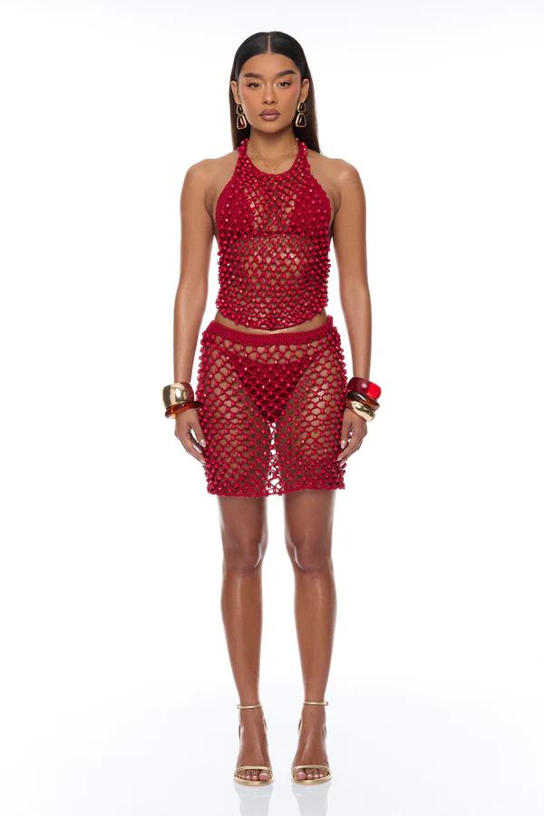 Woman standing in a bronze open-knit crochet set: ruffled long-sleeve top, flared pants, sunglasses, and clutch.