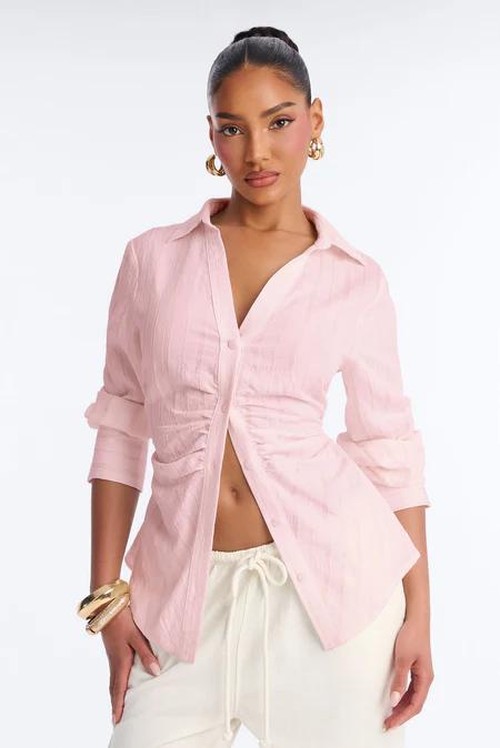 Mujer con camisa rosa abotonada con fruncidos, parcialmente desabrochada, y pantalones crema con cordón. Lleva aros y pulsera dorados.