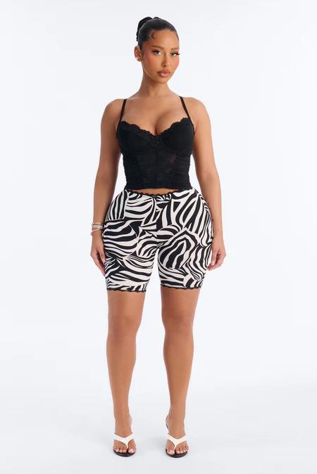Woman standing in a black lace corset top, black and white zebra print biker shorts, and white thong sandals on a white background.