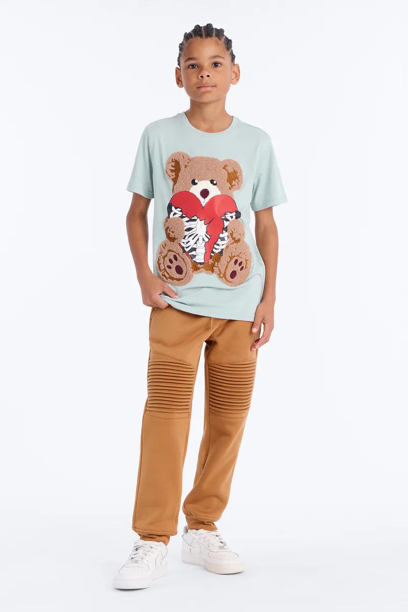 Niño con camiseta gráfica de oso azul claro y corazón roto, joggers marrones acanalados. Moda y ropa juvenil de tendencia.