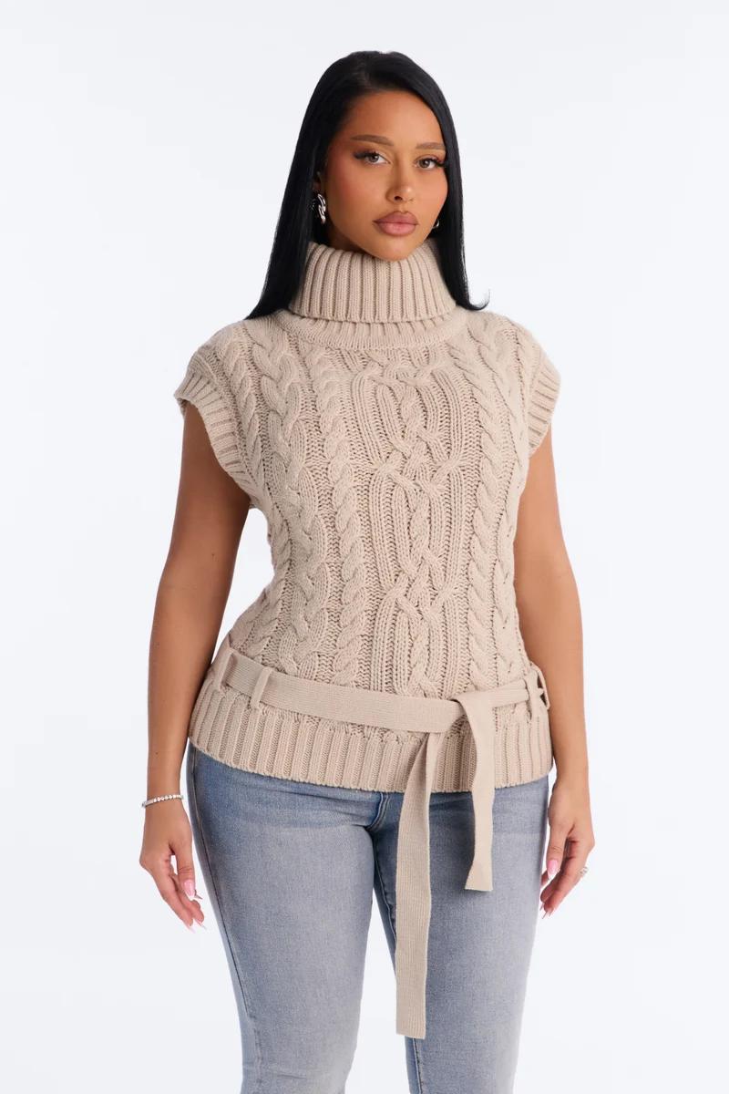 Woman wearing a beige cable-knit sleeveless turtleneck sweater vest with a matching waist belt and jeans.