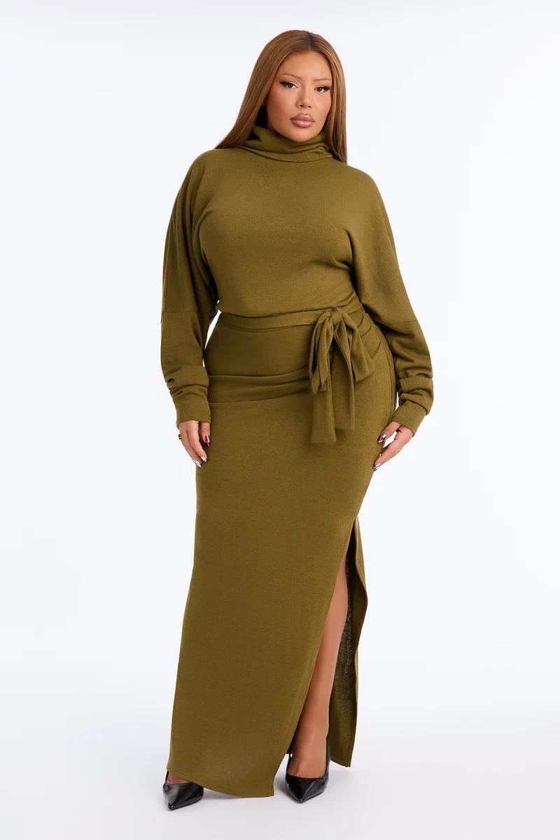 Plus size model poses in an olive green turtleneck knit maxi dress with a tied waist and side slit for a cozy fall fashion collection.