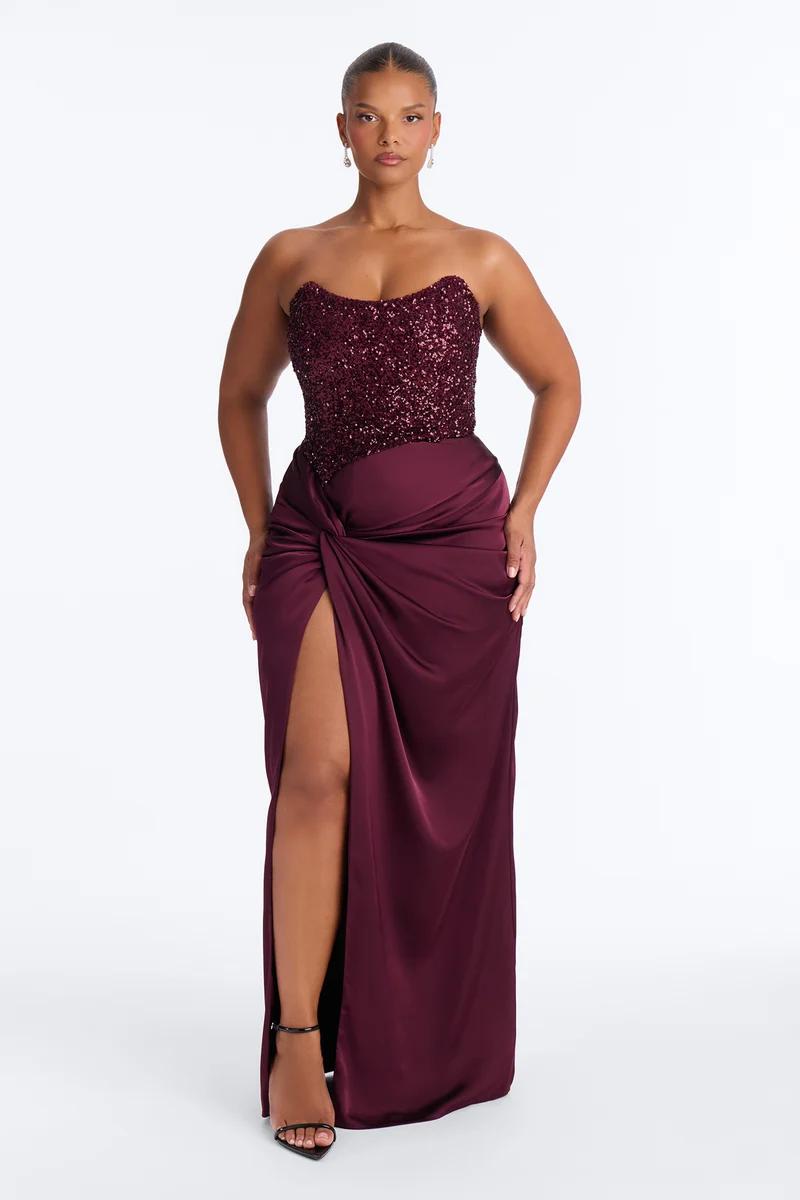 Model wearing a strapless burgundy sequin corset evening gown with a draped satin high-slit skirt, showcasing formal evening wear collection.