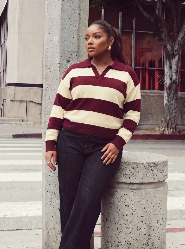 Mujer de talla grande posa con un suéter de rayas color vino y crema y jeans oscuros, modelando la nueva colección de moda de otoño.