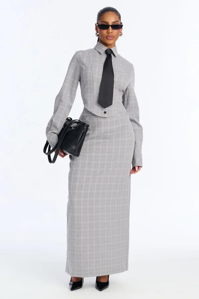 Woman wearing a chic grey plaid maxi skirt and matching long-sleeve top with a black tie and handbag, showcasing modern women's workwear fashion.