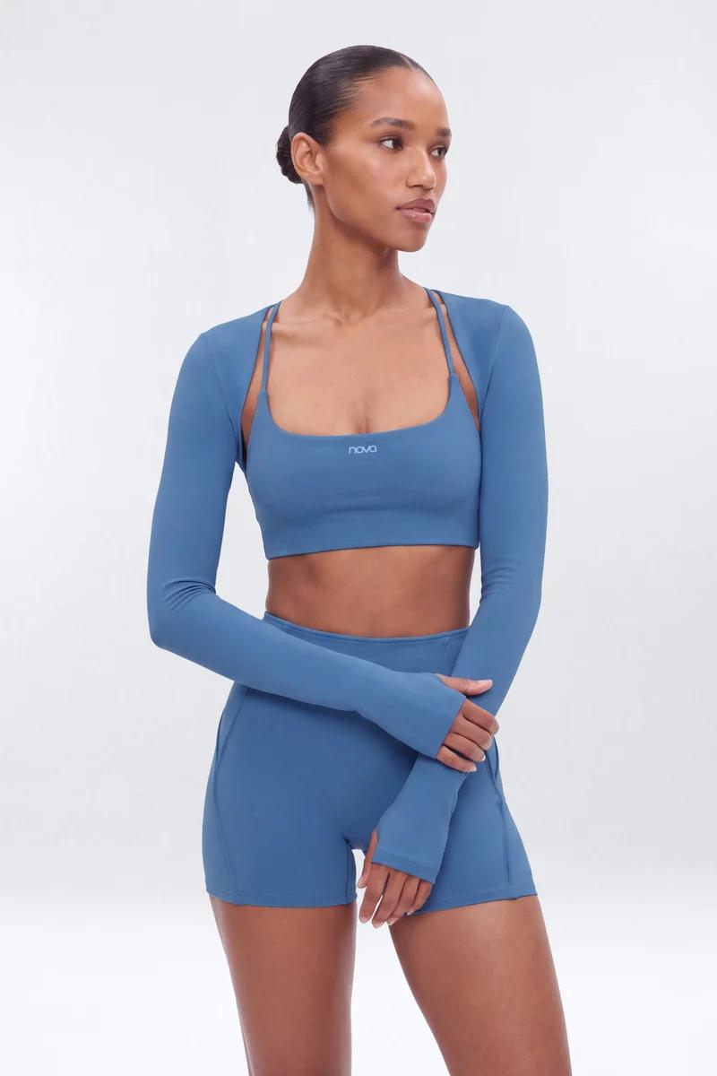 Woman wearing a dusty blue Nova activewear set featuring a sports bra, athletic shorts, and a long-sleeve shrug. Perfect for workout gear.