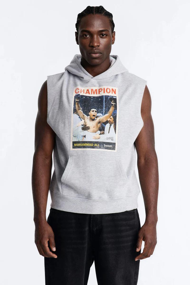 Man wearing a grey sleeveless hoodie with a Muhammad Ali "Champion" boxer graphic and black jeans. Promotes stylish men's casual fashion.