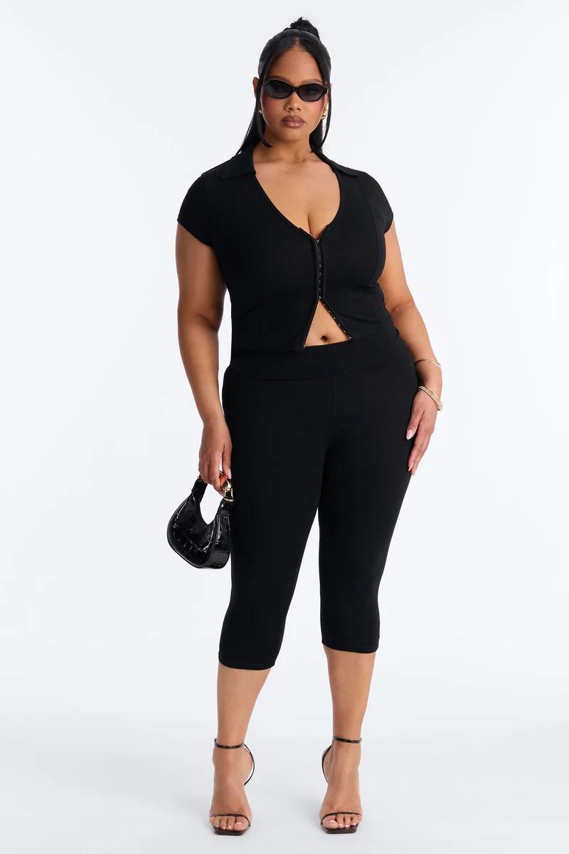 Plus-size model wearing a chic black hook-and-eye short-sleeve top and matching capri pants with heels, showcasing trendy women's fashion.