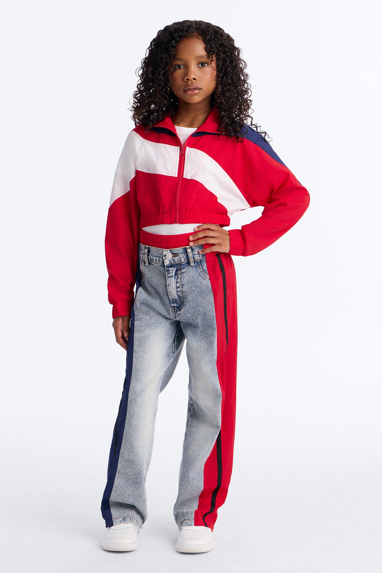 Young girl in a red, white & blue color-block cropped windbreaker & two-tone jeans with side stripes. Kids' fashion.