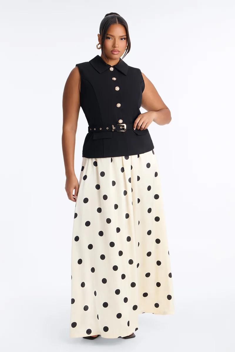 Model wearing a chic plus size outfit featuring a black belted collared vest with gold buttons and a cream polka dot maxi skirt.