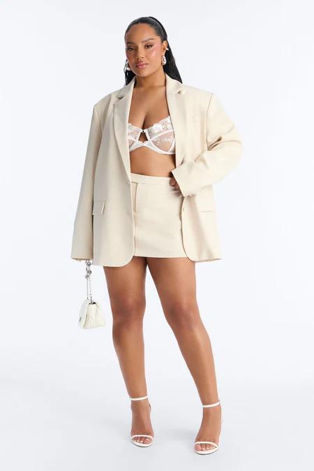 Mujer de moda con blazer crema oversize, minifalda a juego, bralette de encaje blanco, tacones de tiras y bolso acolchado blanco pequeño.