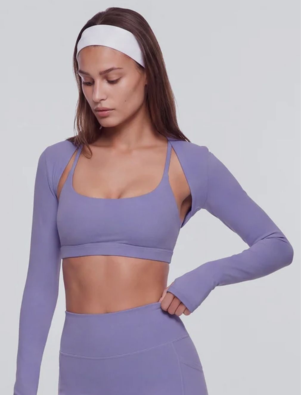 Model wearing a lavender yoga set with a sports bra, matching long-sleeve shrug, leggings, and white headband for a stylish workout look.