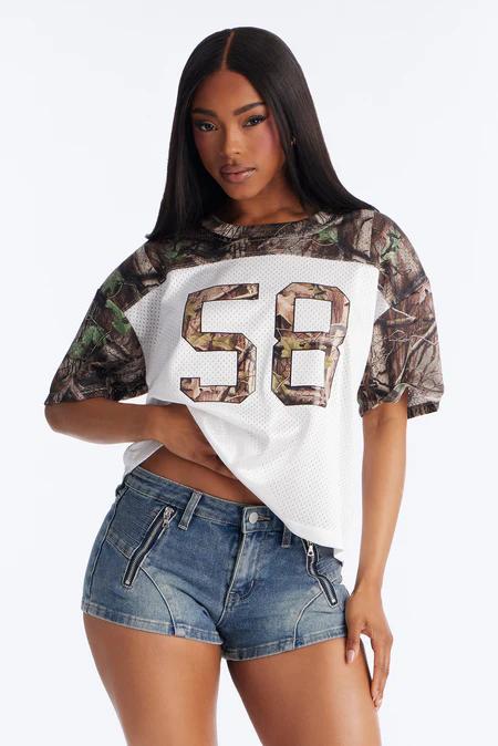 Mujer posando con una camiseta de malla blanca, mangas camuflaje y el número '58', combinada con shorts vaqueros azules con cremalleras.