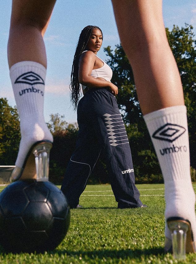 Modelo con top blanco y pantalones Umbro azul marino en campo soleado. Calcetines Umbro, tacones transparentes sobre balón negro exhiben moda deportiva.