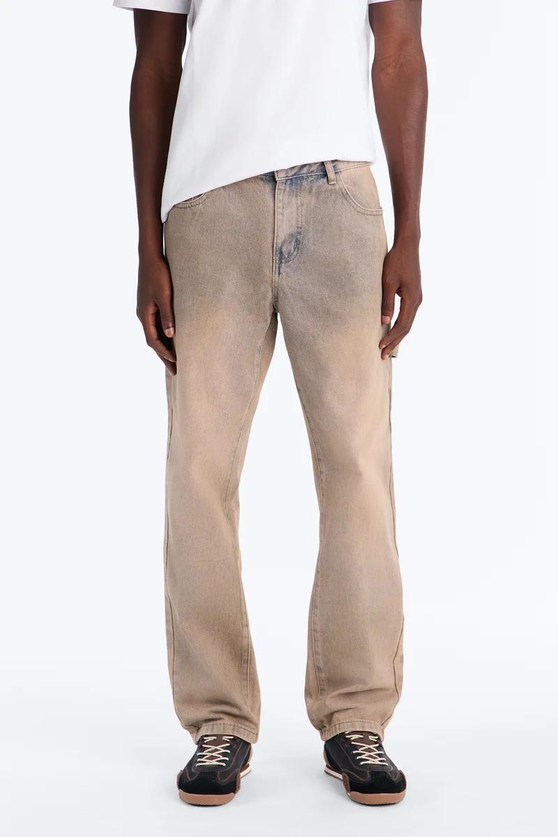 Man in a white tee and stylish faded tan baggy jeans, paired with dark sneakers, showcasing a new casual denim collection.