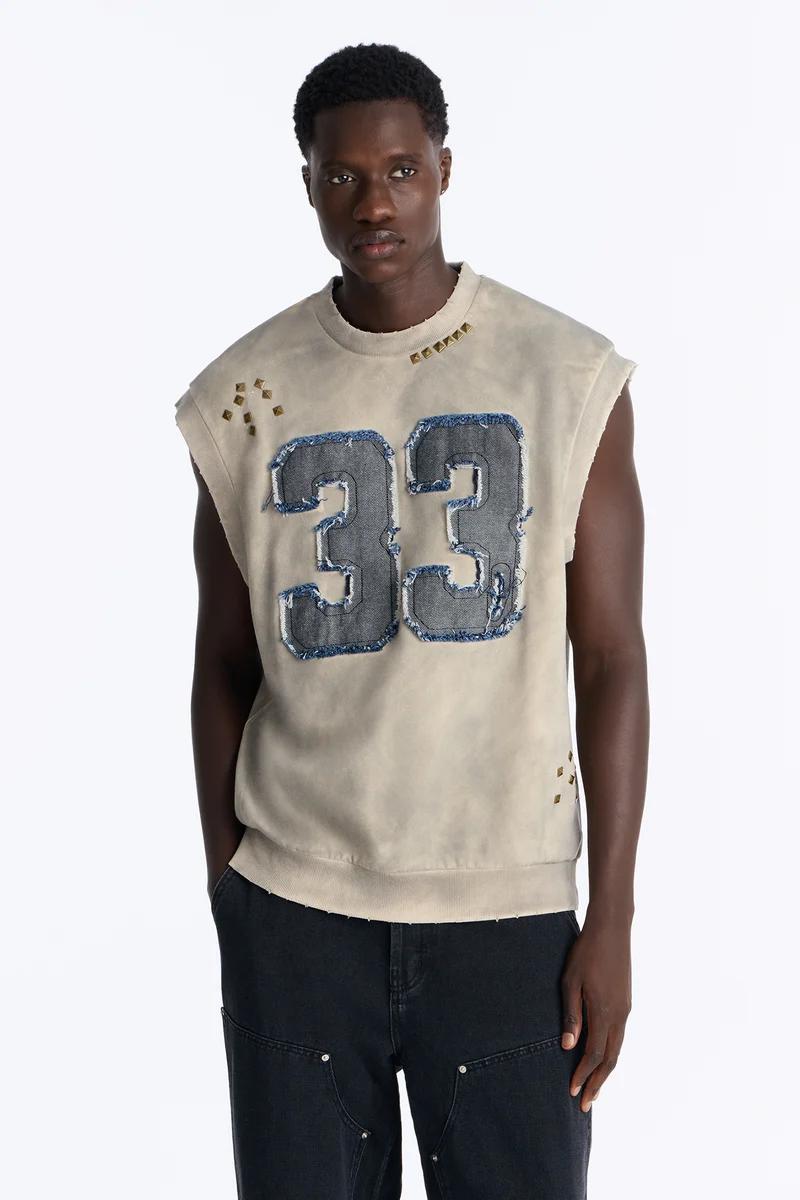 Modern sleeveless beige sweatshirt featuring large distressed denim "33" and pyramid studs, styled with black jeans. Discover new streetwear.