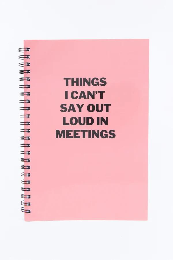 Pink spiral notebook with black wire binding and bold black text: 'THINGS I CAN'T SAY OUT LOUD IN MEETINGS' on its cover.