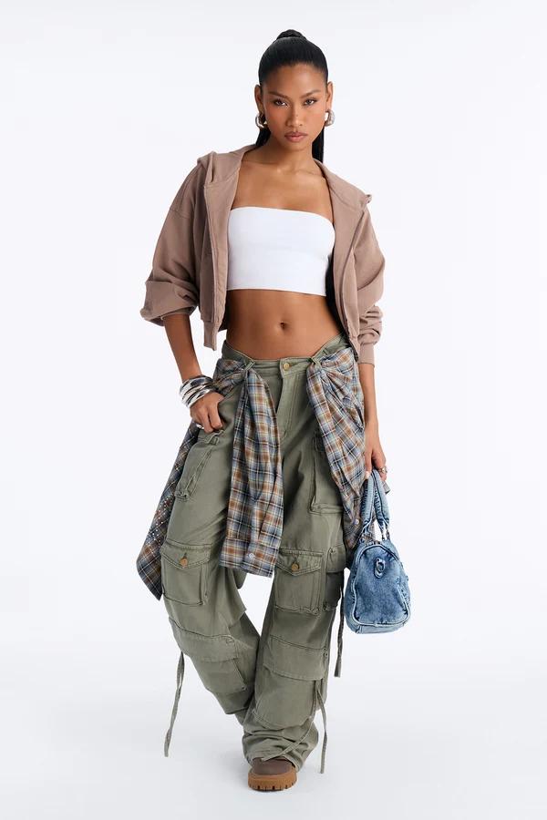 Modelo con estilo streetwear Y2K vistiendo pantalones cargo verdes, top blanco, sudadera marrón y camisa de cuadros para la nueva colección.