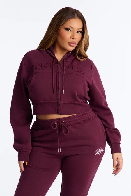 Persona sonriente vistiendo sudadera corta granate con cremallera, texto en relieve y pantalones a juego, posando sobre fondo blanco.