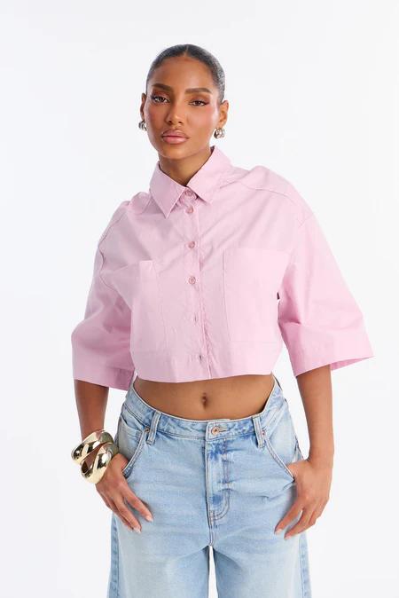 Mujer elegante con camisa corta rosa claro de botones, jeans de lavado claro, pendientes de aro plateados y pulseras doradas, posando sobre fondo blanco.