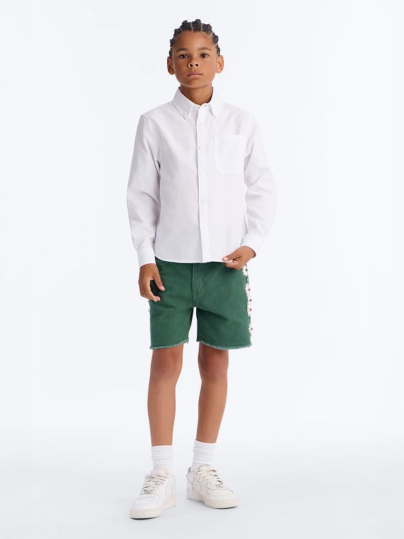 Young boy models a crisp white button-up shirt, green frayed shorts with embroidery, and sneakers, showcasing stylish kids' casual wear.