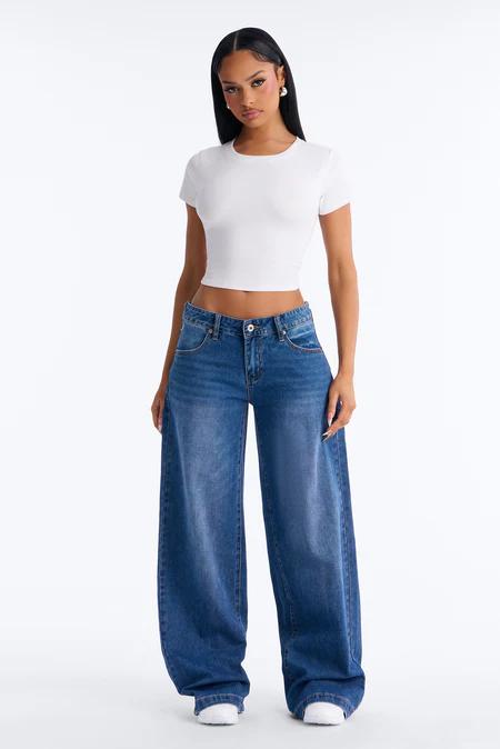A woman wearing a white cropped t-shirt, loose blue wide-leg jeans, and white sneakers stands facing forward on a plain background.