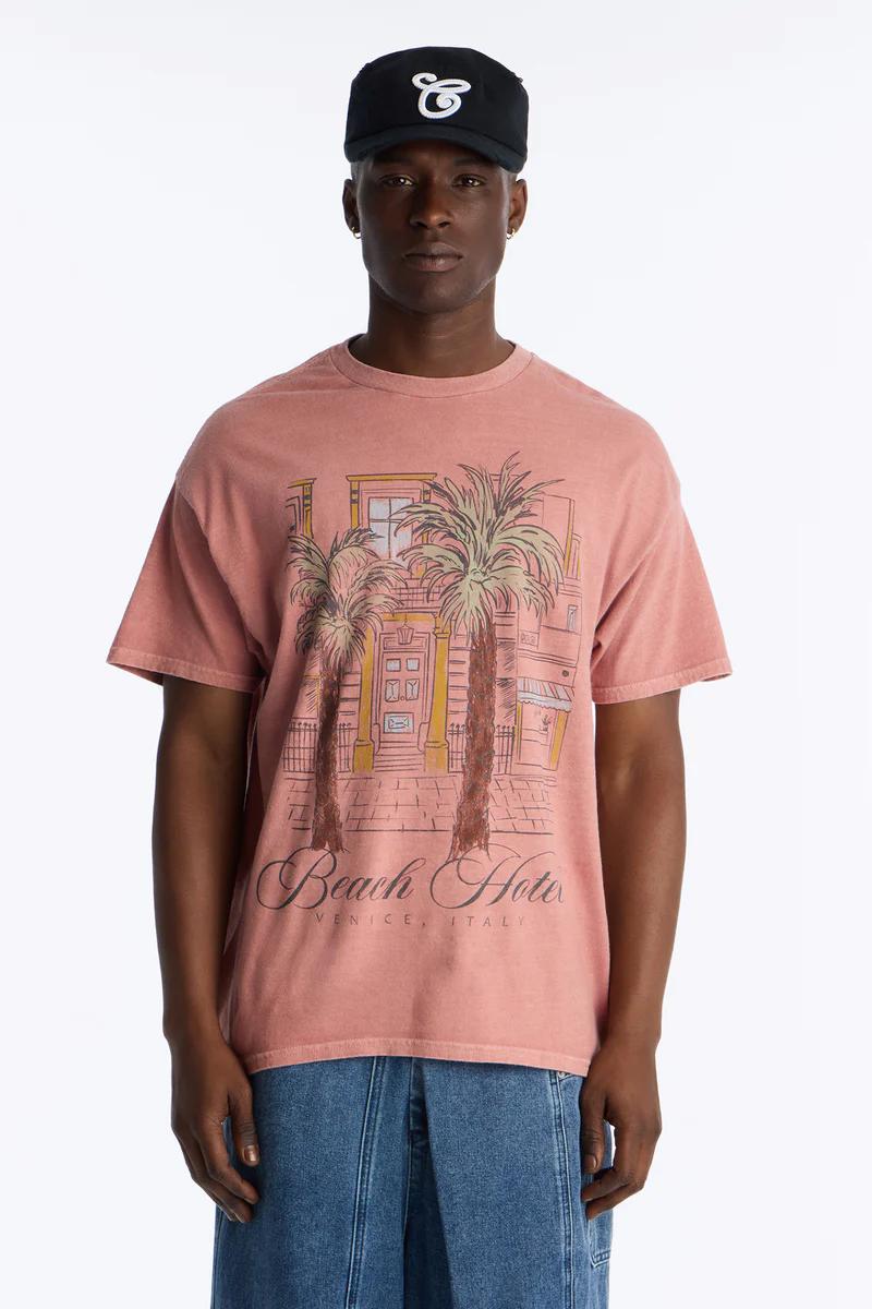 Men's faded pink graphic tee showcasing 'Beach Hotel Venice, Italy' and palm trees, styled with a black cap and jeans.