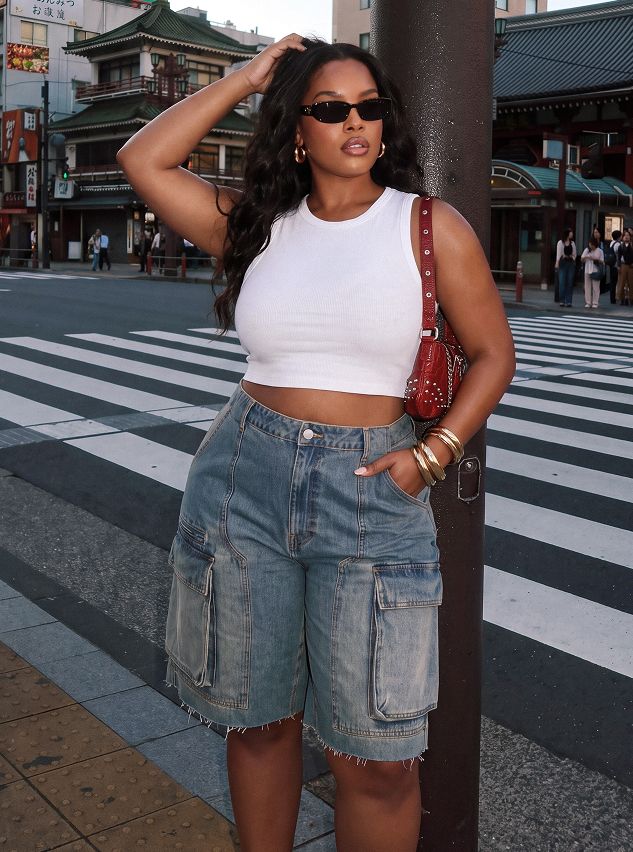 Stylish woman wears a white ribbed crop top, light wash denim cargo shorts, red bag & sunglasses on a city street, showcasing summer fashion.