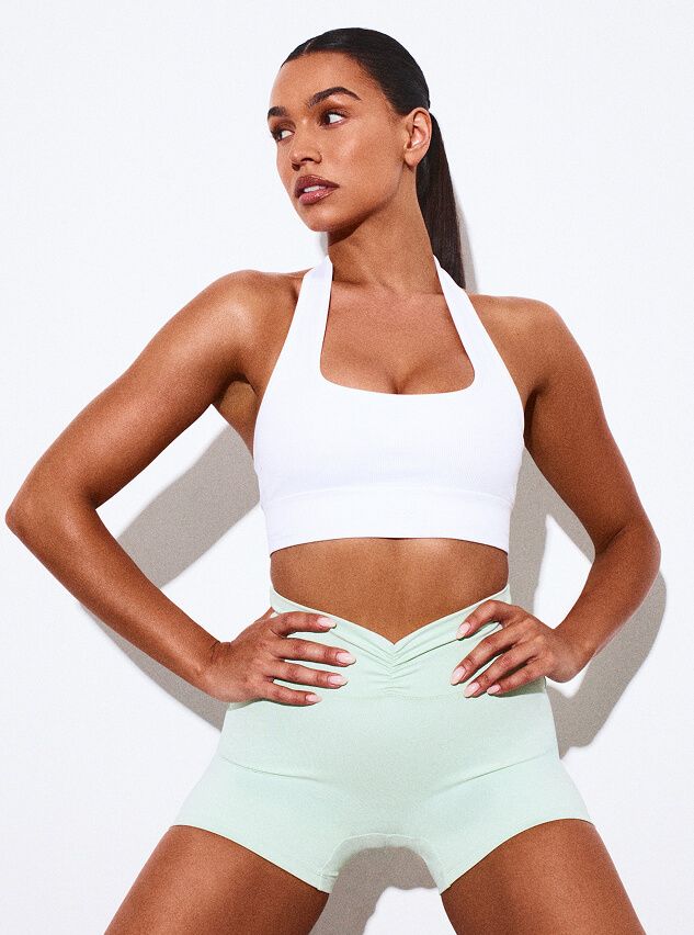 Woman modeling a white halter neck sports bra and mint green ruched active shorts. Showcasing new fitness wear collection.