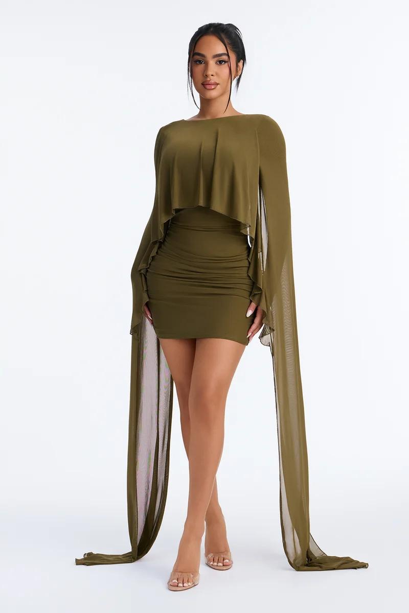 Woman wears an olive green bodycon mini dress featuring dramatic floor-length cape sleeves, showcasing a trendy evening fashion line.