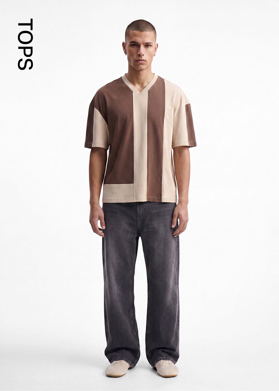 Hombre modela camiseta color block marrón y beige con cuello V, jeans carbón y zapatillas, mostrando tops masculinos de moda.