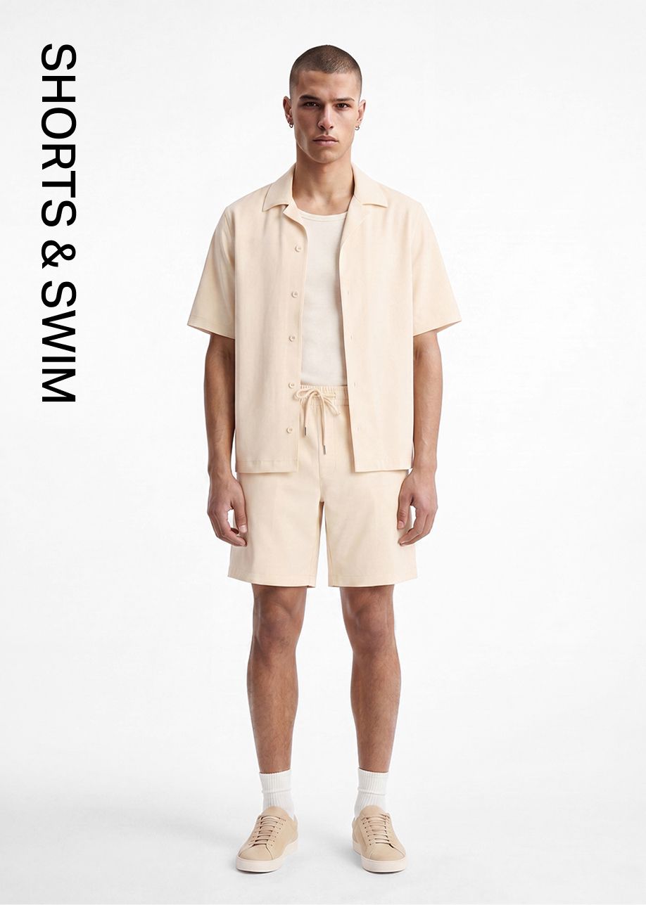 Man in light beige short-sleeve shirt, t-shirt, and matching drawstring shorts with sneakers, showcasing men's casual shorts and swim styles.