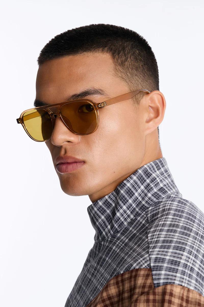 Man models trendy brown aviator sunglasses with vibrant yellow lenses, showcasing men's eyewear style. Wears a grey and brown plaid shirt.