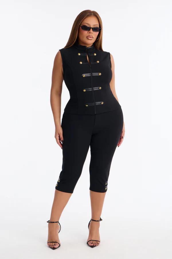Confident woman models a black sleeveless vest with gold buttons, matching capri pants, and heels, showcasing modern fashion.