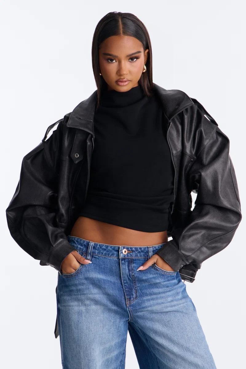 Woman in oversized black leather bomber jacket, black mock neck crop top, and blue jeans.