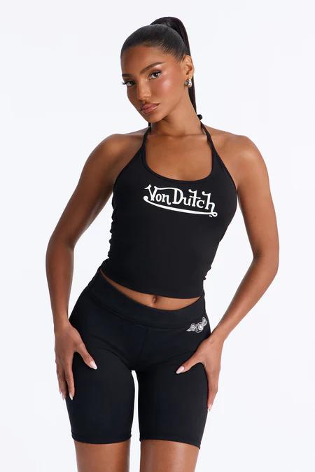 Mujer con top halter negro con logo blanco de Von Dutch y shorts ciclistas negros con diseño alado sutil, posando con confianza.
