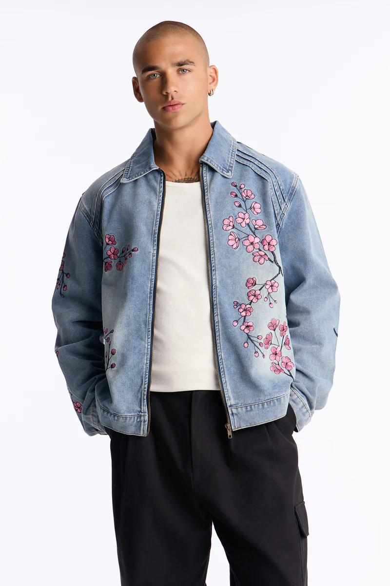 Man modeling a stylish light blue denim jacket with intricate pink cherry blossom embroidery, perfect for a new collection.
