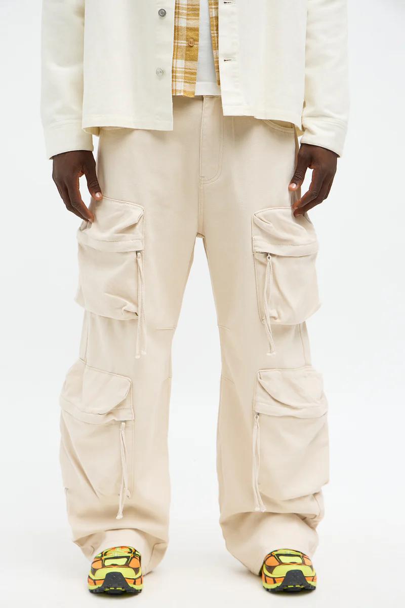 Modelo con pantalones cargo beige holgados y bolsillos, chaqueta blanca, camisa de cuadros y zapatillas vibrantes, promocionando moda casual masculina.