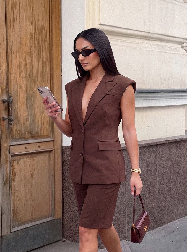 Stylish woman in a brown sleeveless blazer and matching shorts suit, checking phone. Perfect for a chic summer fashion look.